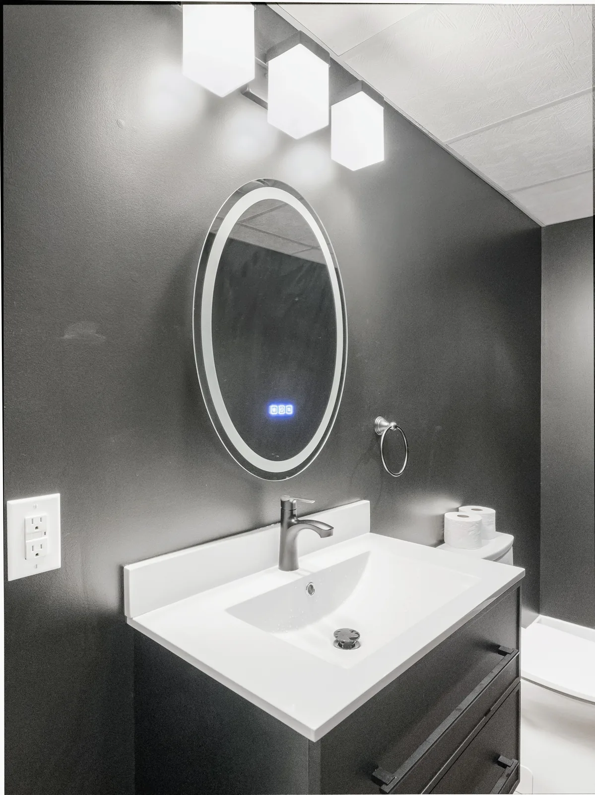 Basement bathroom vanity — white basin on a black wall-hung unit under a backlit oval mirror