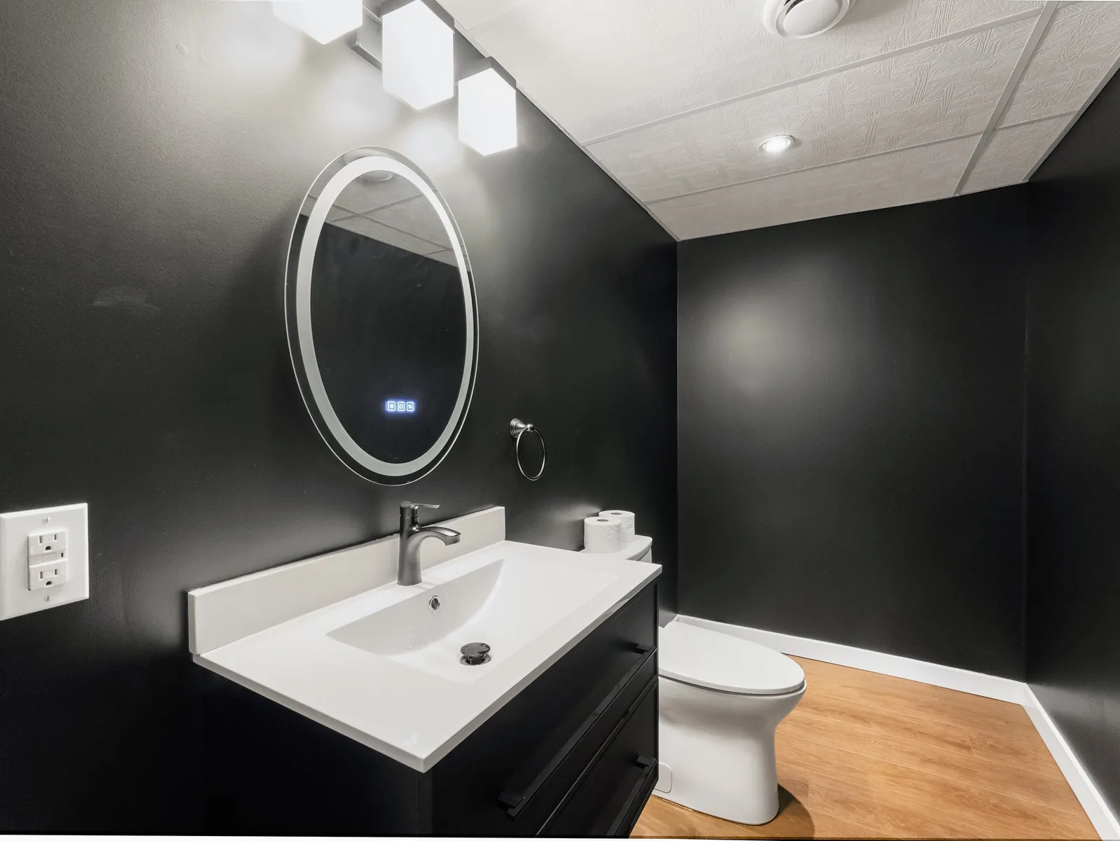 The bathroom in full, charcoal walls with square glass sconces over the mirror