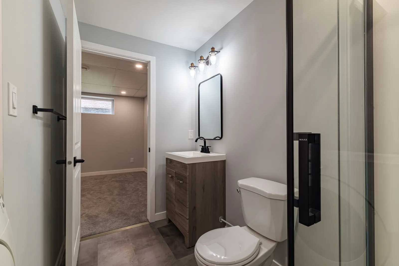 Basement bathroom from the door, black fittings and a glass shower, the carpeted rec room beyond