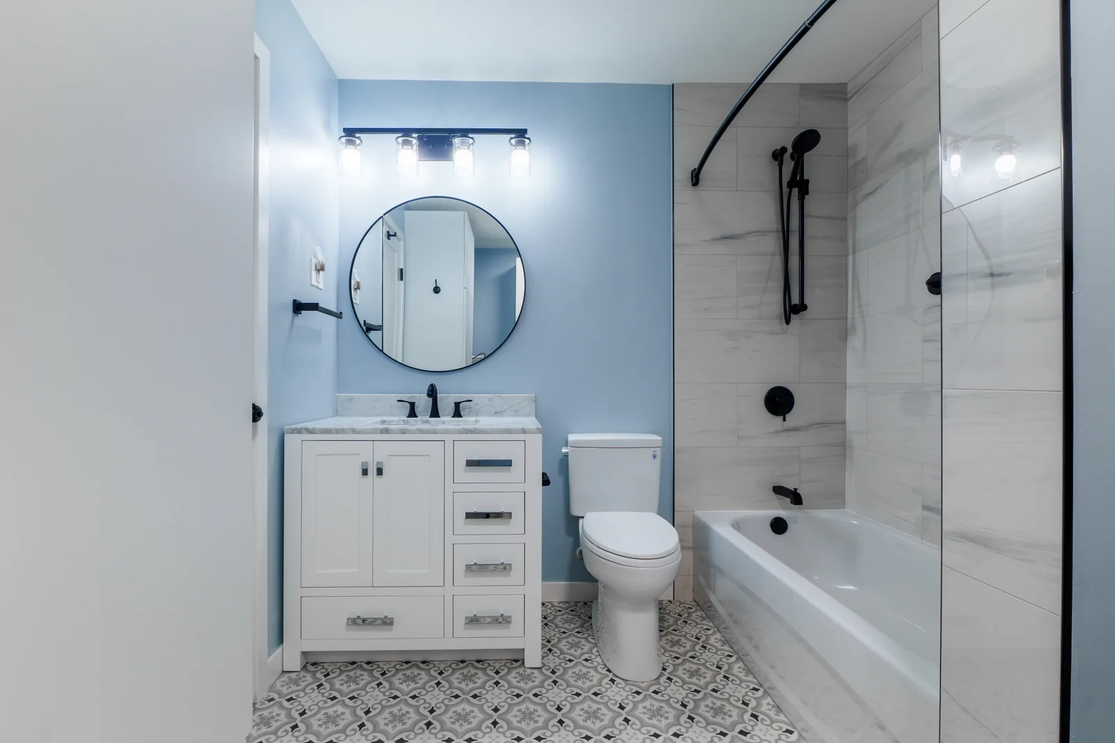 Pale blue bathroom with a white shaker vanity, round black mirror and a marble-look tub surround