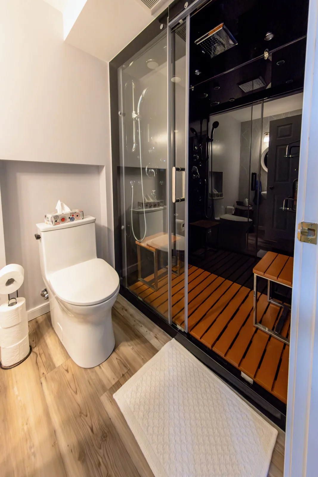 Basement bathroom with a black-glass shower cabin, teak floor inside it, and a white toilet in a recessed alcove