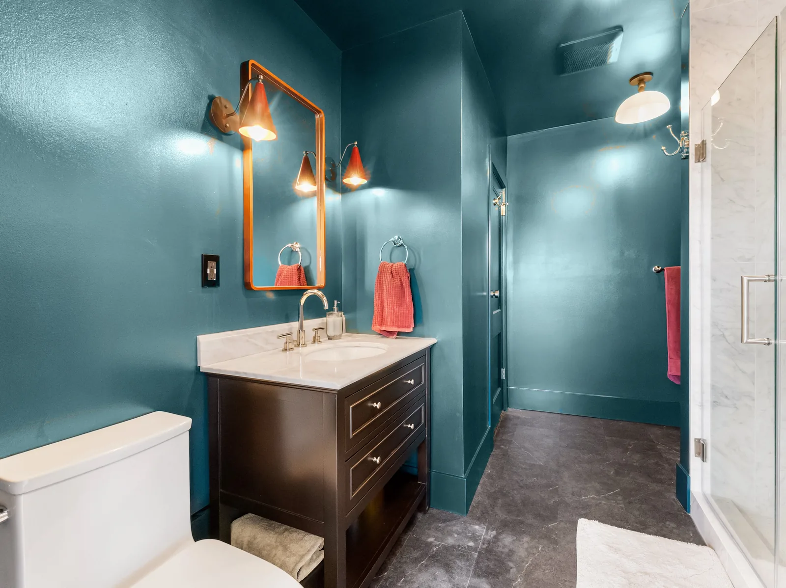 Teal bathroom end to end — dark wood vanity under a brass mirror, marble-look shower beyond