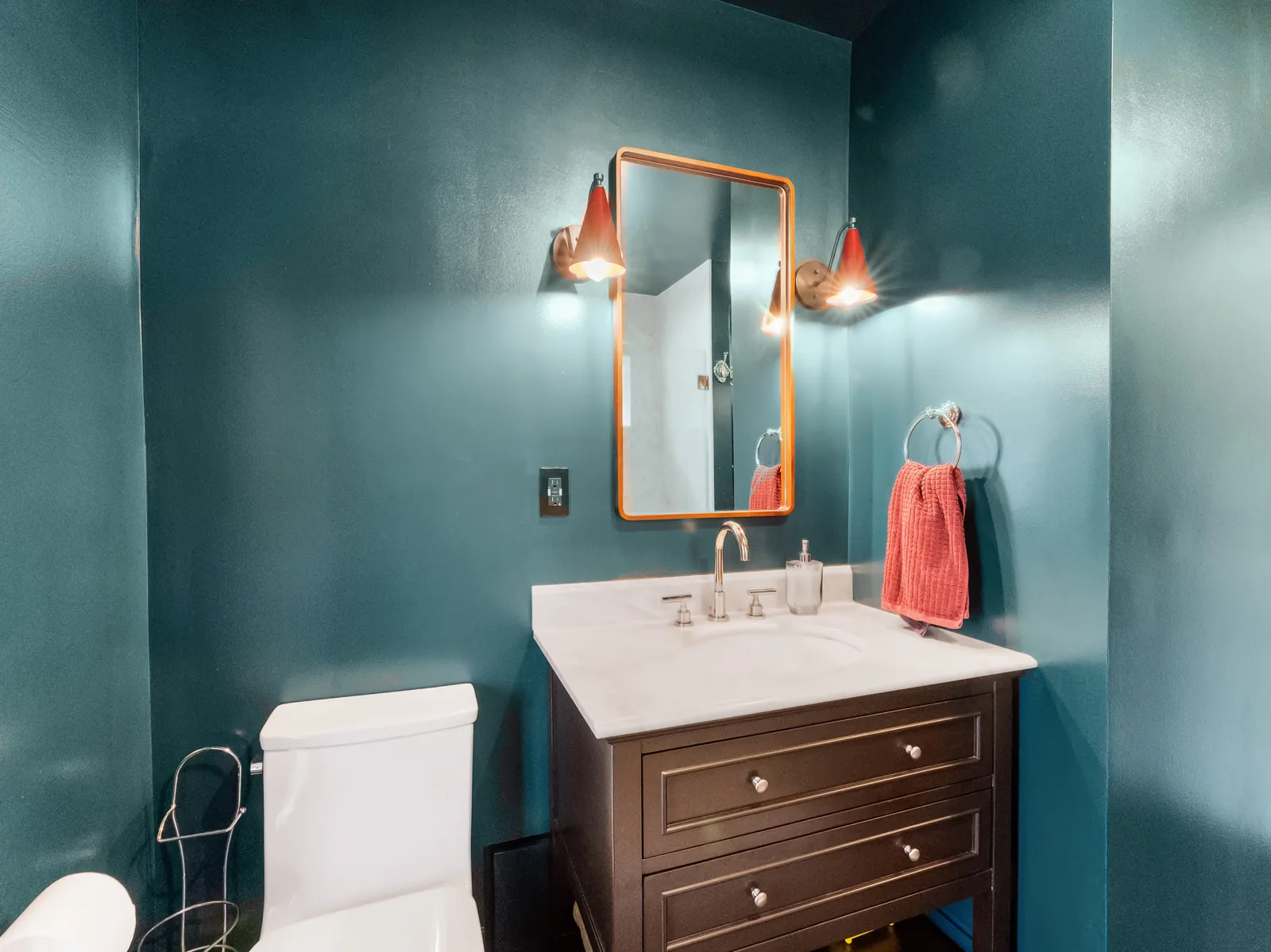 Dark wood vanity with a white stone top, brass-framed mirror and copper sconces on deep teal