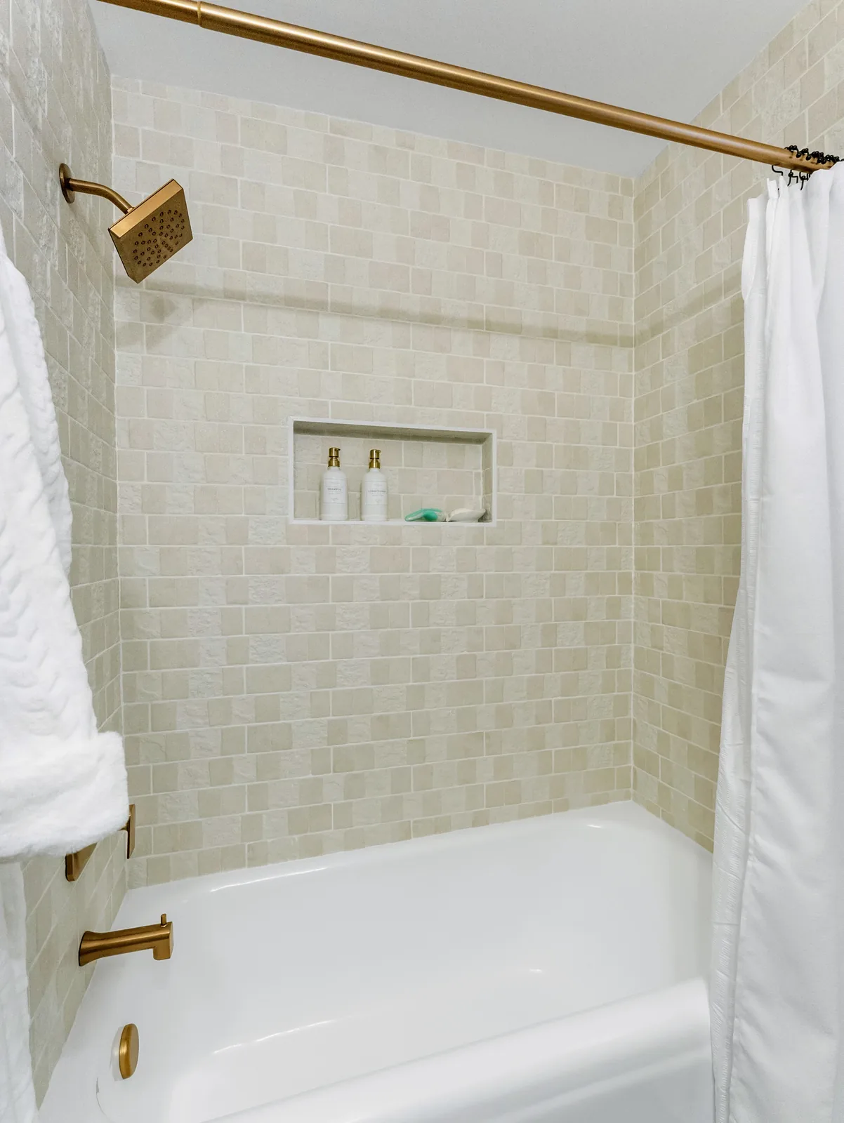 Tub-shower with a textured white tile surround, recessed niche and a brass rail