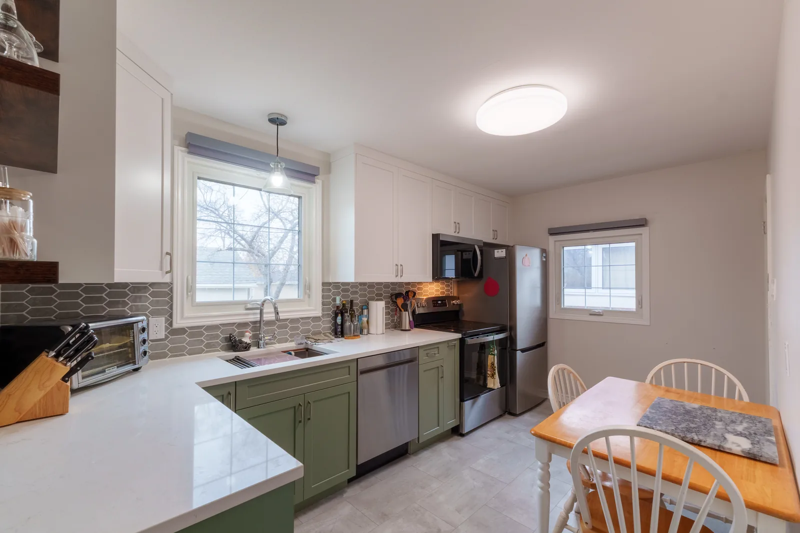 Kitchen in sage-green base cabinets and white uppers, quartz counters and a mosaic backsplash