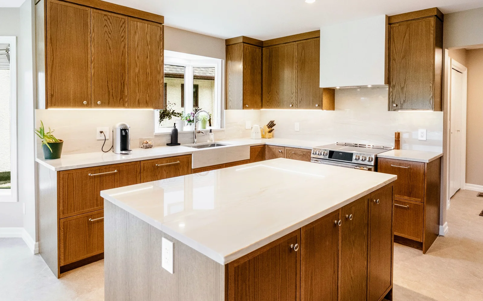 After — the same kitchen rebuilt: stained-oak cabinetry to the ceiling, a marble-veined quartz island, an apron sink and a panelled hood over the range