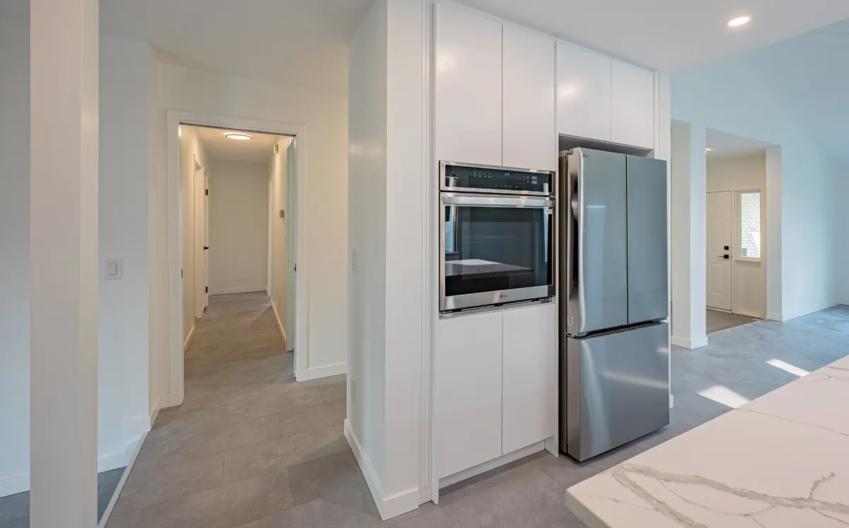Wall oven and stainless fridge set into a tall white cabinet run, the hallway beside it